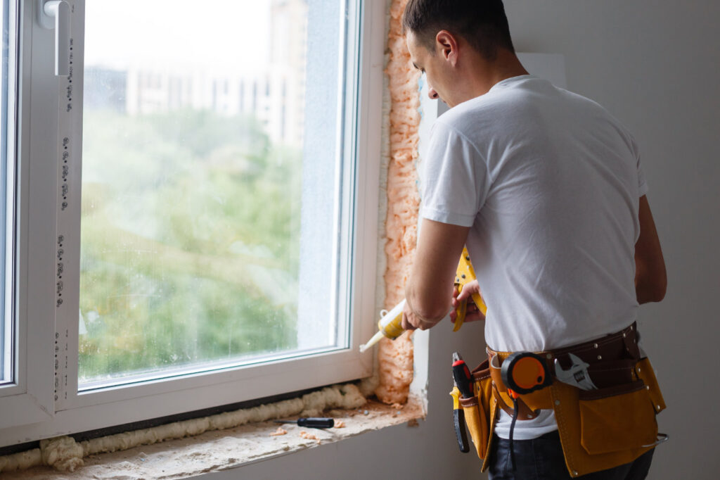 Worker caulking and air-sealing a window frame filled with expanding spray foam insulation.