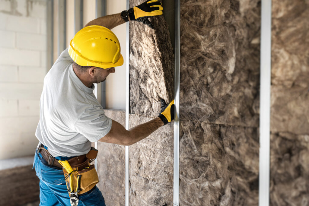 Worker in hard hat fitting mineral wool batt insulation between metal wall studs.