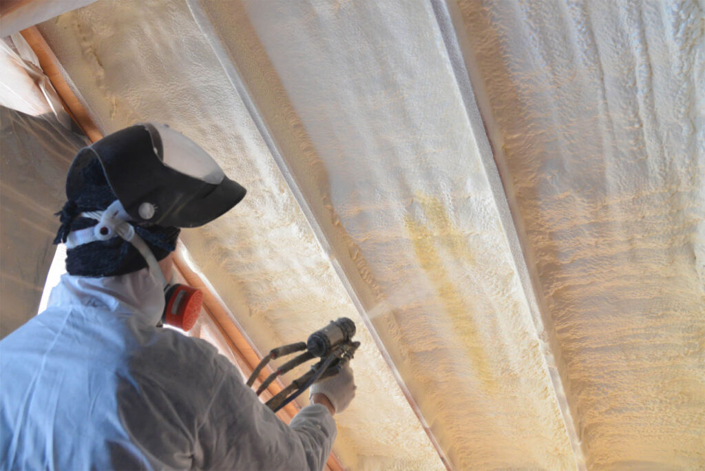 Worker sprays foam insulation between roof joists in an attic ceiling