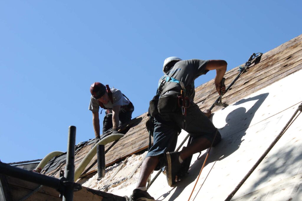 Two harnessed workers blowing cellulose insulation into an open roof deck under blue sky.