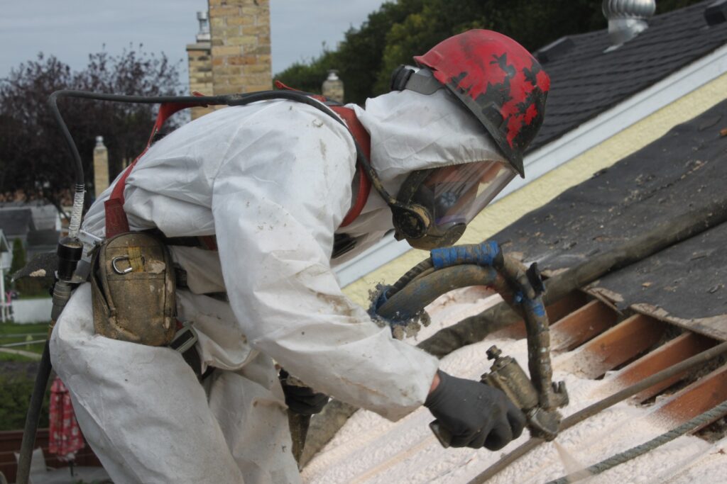 Worker in white protective suit and respirator spray-foam insulating an open roof deck.