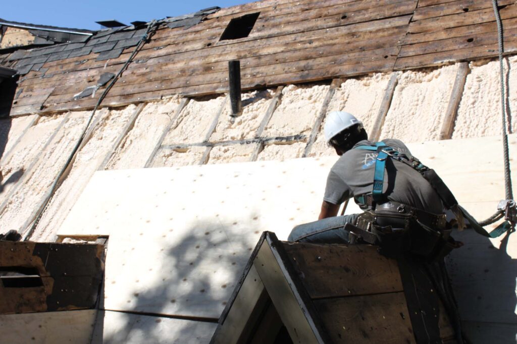 Roofer in harness on stripped roof deck with exposed spray-foam insulation and new plywood sheathing.
