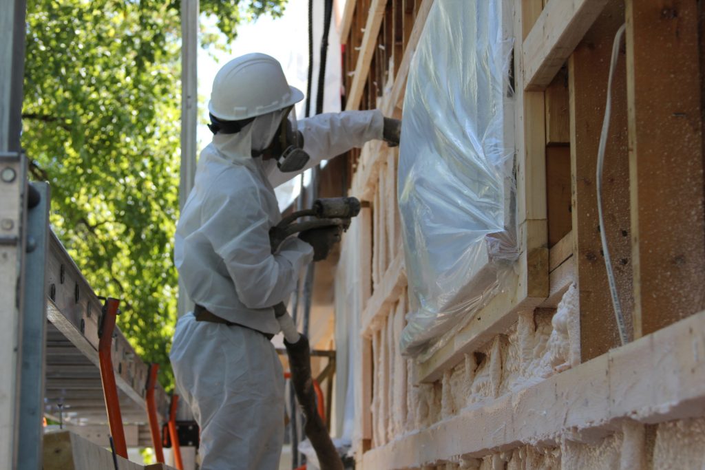 Worker in protective gear spraying foam insulation onto the exterior wall of a house under construction, with a window covered in plastic sheeting for protection.